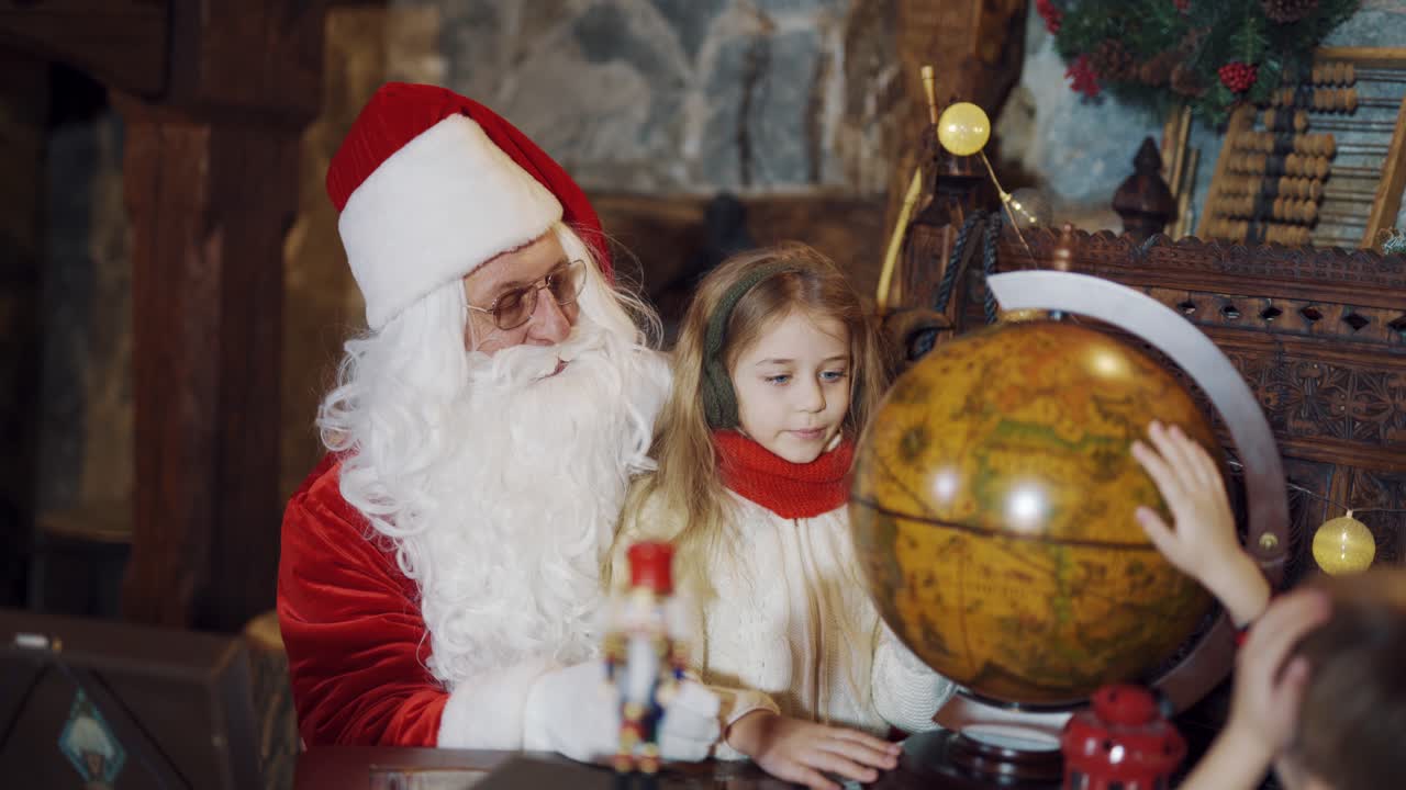 Santa Claus shows little girl a globe. Santa in red costume with white beard sitting in the room with child and telling about the important places on a big globe. Christmas time.
