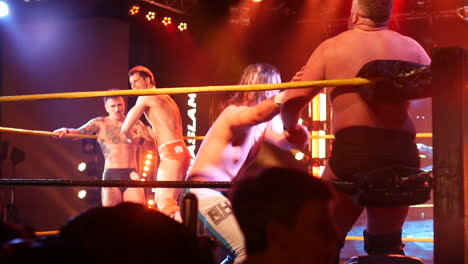 four pro wrestlers engage in a tense theatrics in the wrestling ring, with one grappling an opponent near the corner ropes.