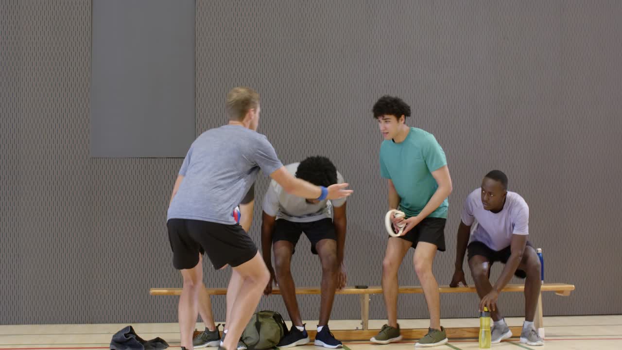 Diverse Männer lösen eine Teambesprechung auf, sitzen auf der Bank im Sportoutfit; ein kniender Mann hält einen Ball und gibt Feedback.