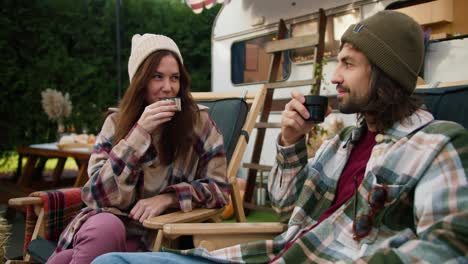 a happy brunette girl in a white cap and a checkered shirt drinks tea with her brunette boyfriend in a green checkered shirt sitting on special chairs during a picnic near her trailer in the forest in the summer