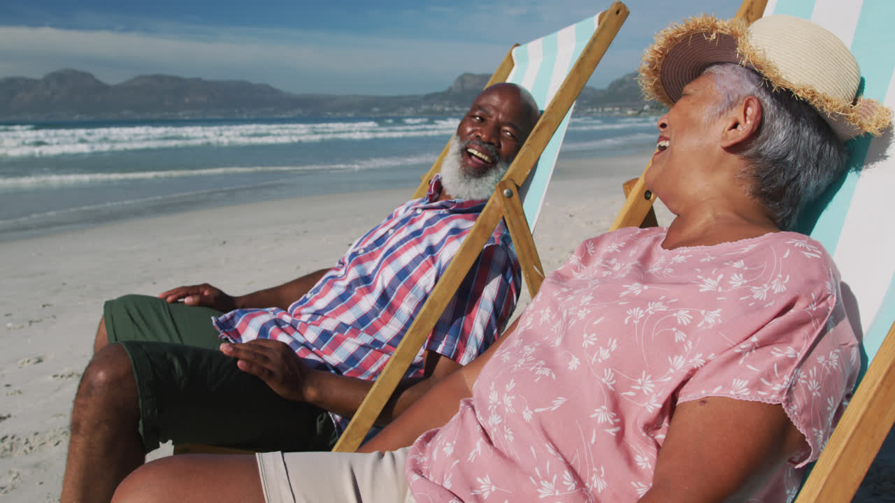 una pareja de ancianos afroamericanos sentados en camas de sol y sonriendo en la playa