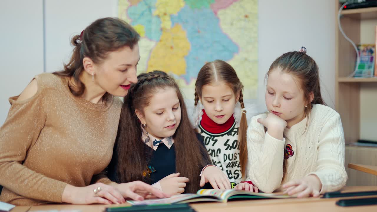 Elementary schoolchildren and a teacher. Little girls sitting at the table with teacher and looking into a book together. Learning process at school.