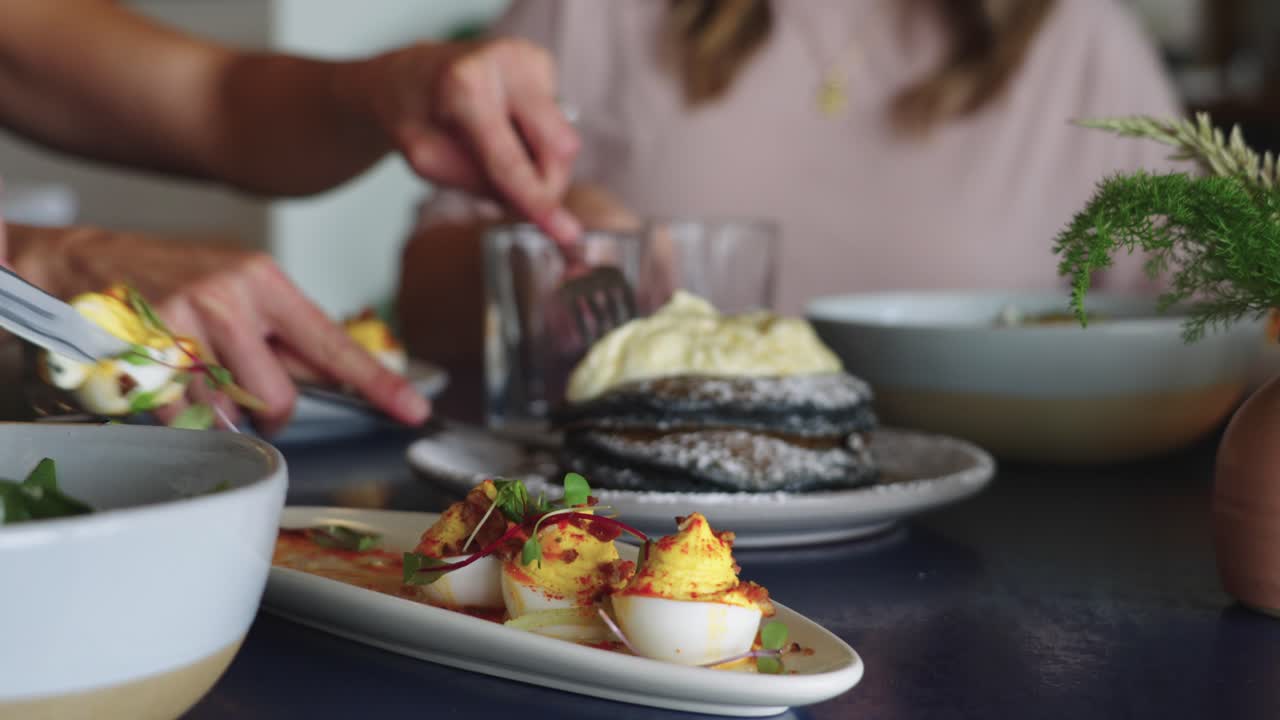 Video 4K di tre donne che gustano pancake di mais blu guarniti con panna e uova ripiene con microgreens, durante un elegante brunch con cibo gourmet e convivialità.