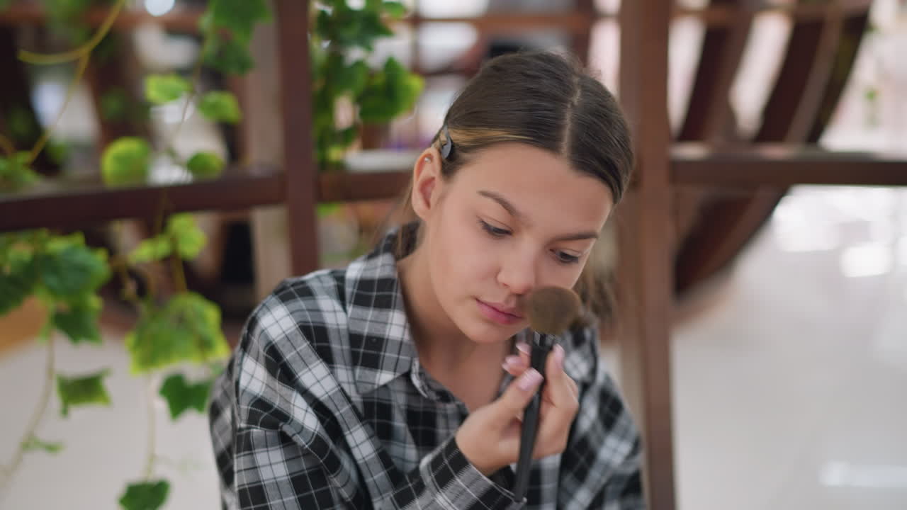 joven dama empapando su nariz con pincel de maquillaje frente al espejo mientras se aplica maquillaje, varios productos cosméticos son visibles en la mesa, destacando su rutina de belleza
