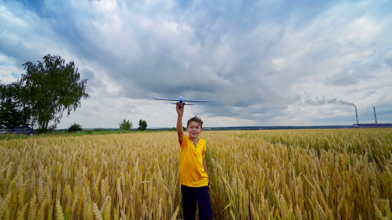 Little boy on yellow field. Happy child playing outdoors in summer. Kid holds a toy plane and imitate flight over the meadow. Front view.