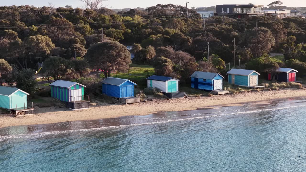 Drone pans over vibrant beach huts, sandy shoreline, and calm water in soft daylight