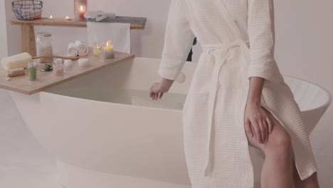 woman in bathrobe sitting on edge of bathtub while touching water