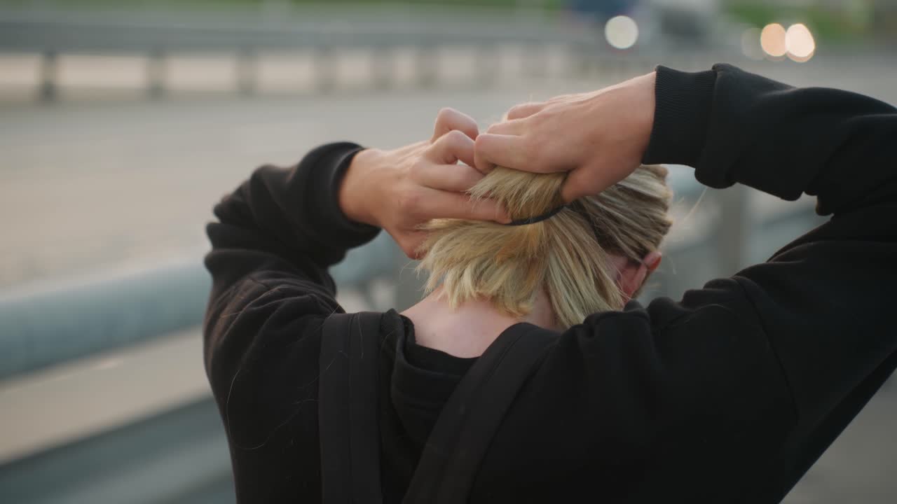 Blonde vrouw die staart maakt op snelweg, forens verandert in zelfverzekerde reiziger, handen gladde vlechten en strakgetrokken elastiek, gloeiende skyline, vangrail en passerende auto's, klaar om te vertrekken