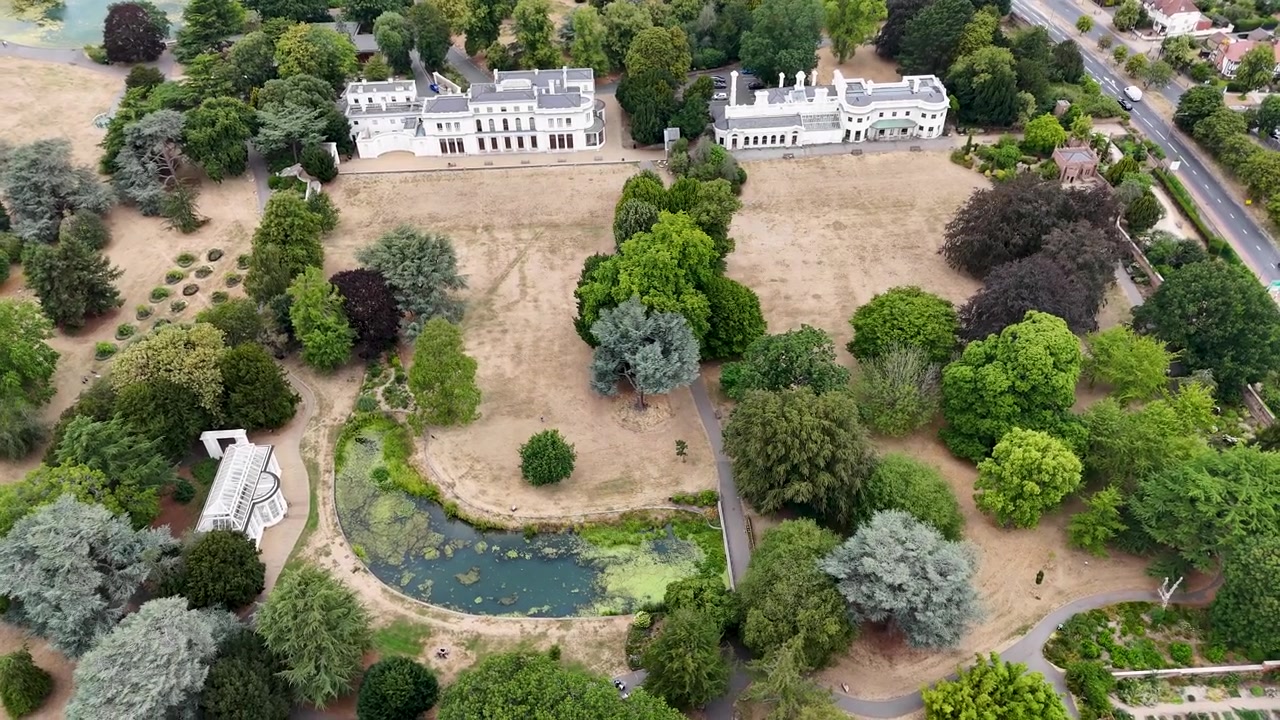 Deux manoirs en stuc blanc se dressent dans un parc historique avec un étang ornemental, l'herbe brûlée par la canicule.
