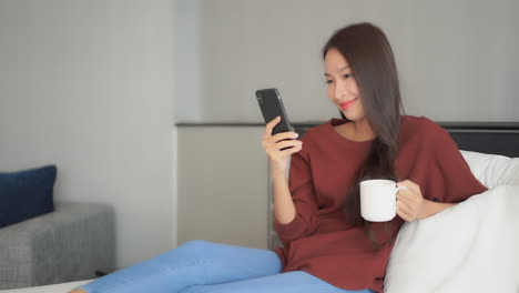 asian woman with a beautiful smile is using her mobile phone holding a cellphone in one hand and typing on the screen, holding a cup on morning coffee