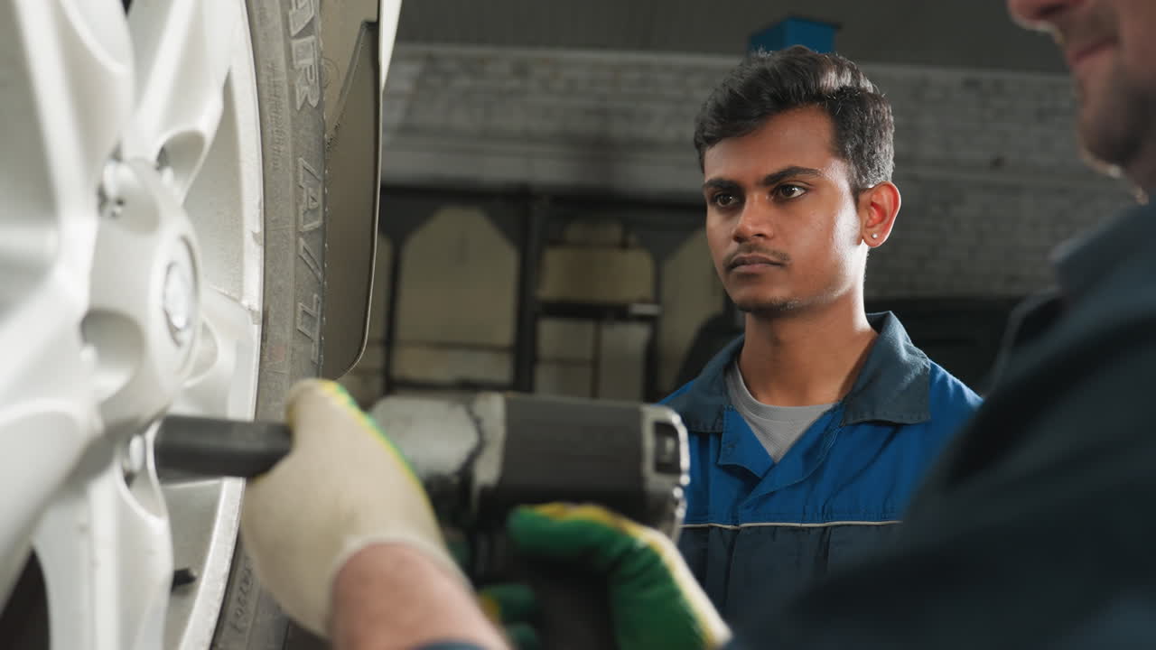 primer plano de una mano con guantes usando una herramienta neumática para apretar los pernos de la rueda de un automóvil en un garaje, un colega está ligeramente borroso en el fondo, observando atentamente el proceso de reparación