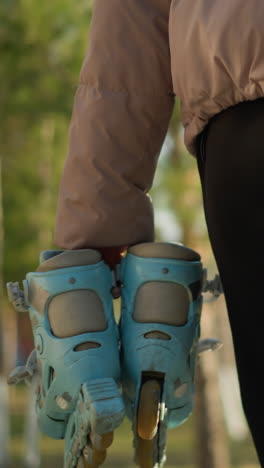 primer plano de una persona con un par de patines, con pantalones negros y una chaqueta de melocotón, caminando por un parque con un fondo borroso de árboles