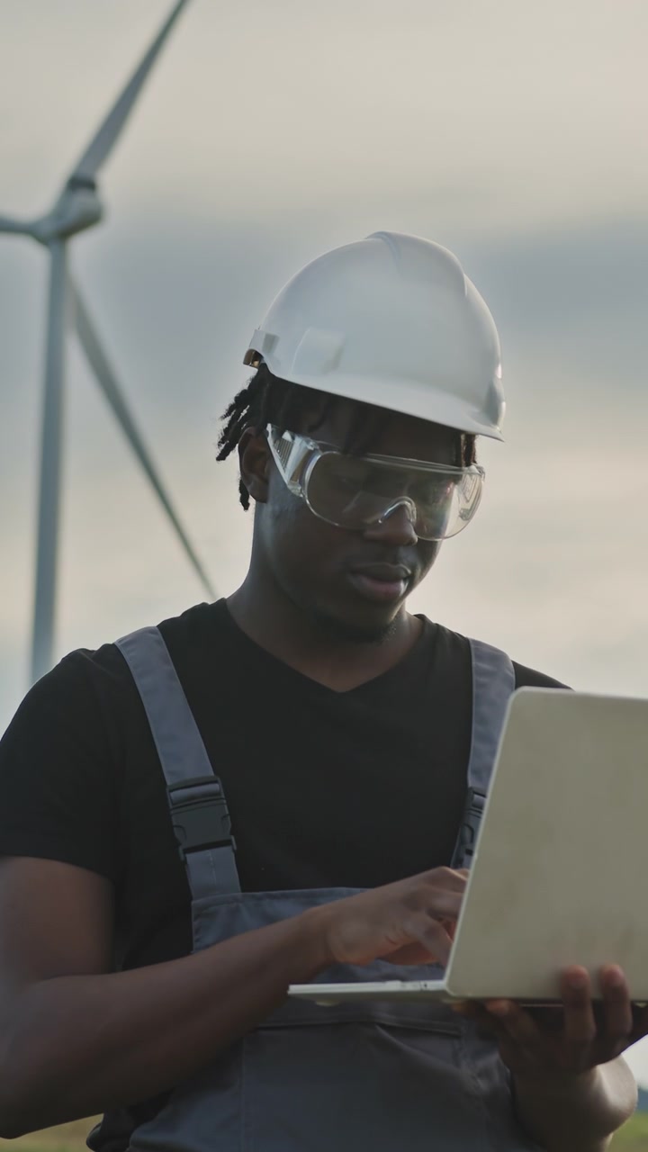 Engineer in hard hat and safety glasses uses laptop near wind turbines monitoring renewable energy