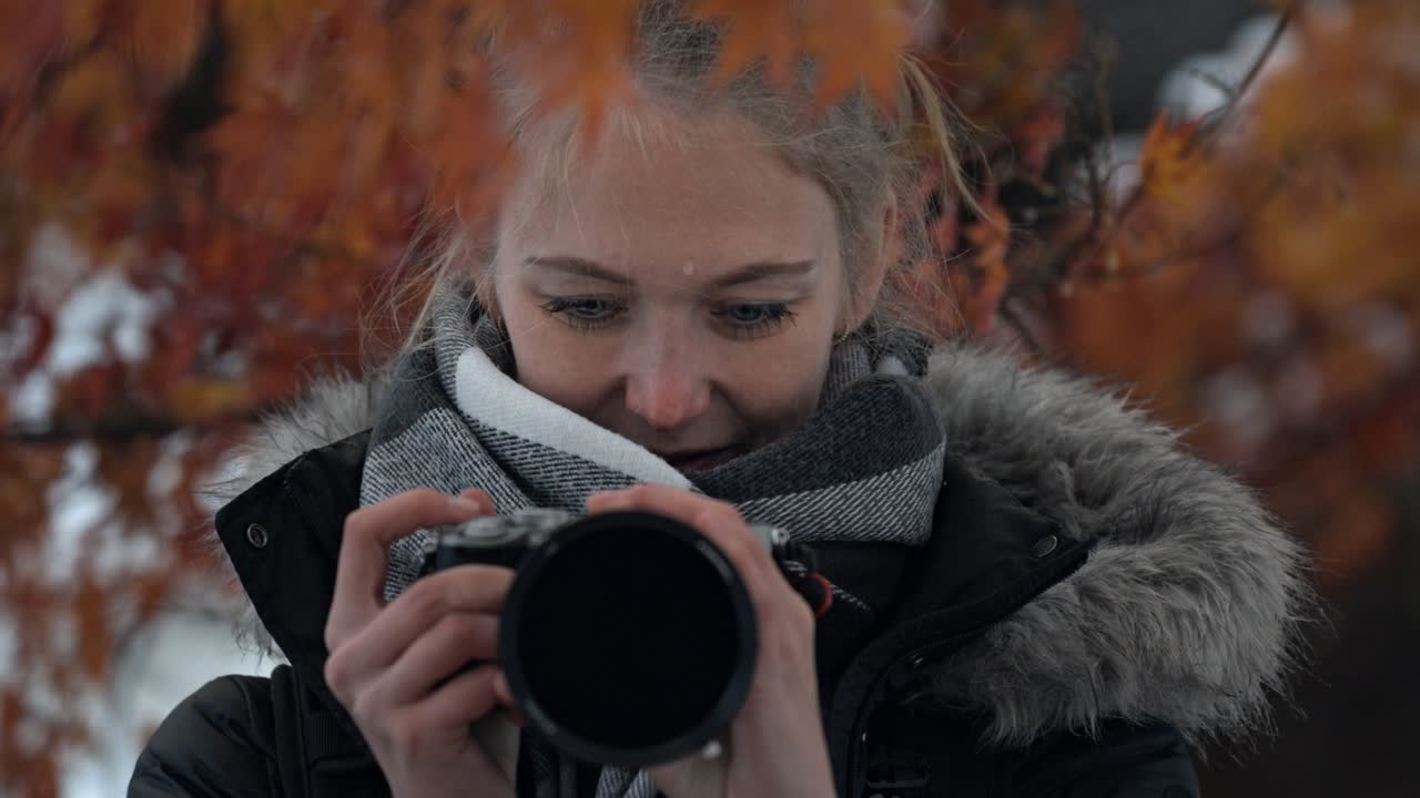 A woman captures the beauty of Shirakawa-go as she stands surrounded by vibrant red autumn leaves, with gentle snowflakes falling around her.