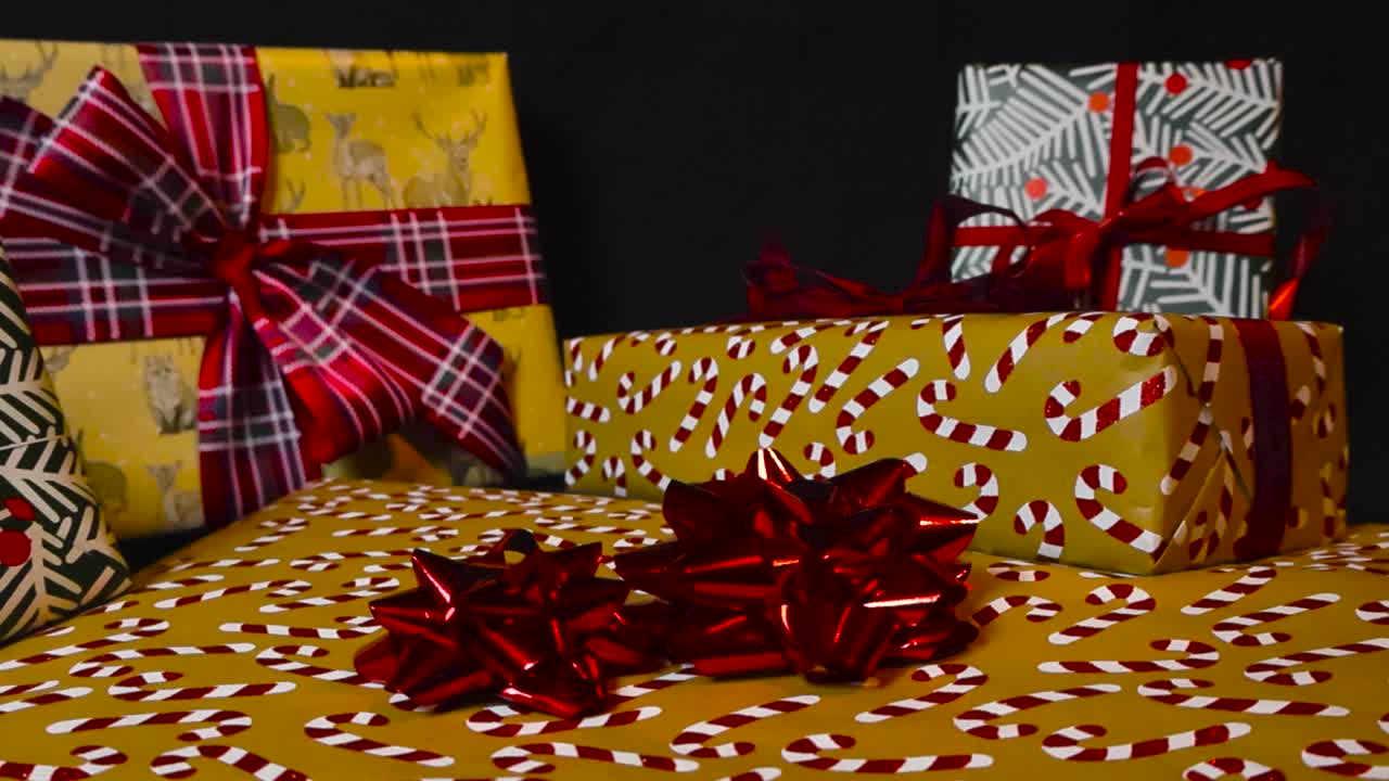 다채롭게 포장된 선물과 크리스마스 선물이 어두운 검은색 배경에 천천히 빨간 리본으로 가로질러 미끄러집니다. 선물은 스튜디오 조명 아래 잘 조명되고 서류에는 사탕통이 있습니다.