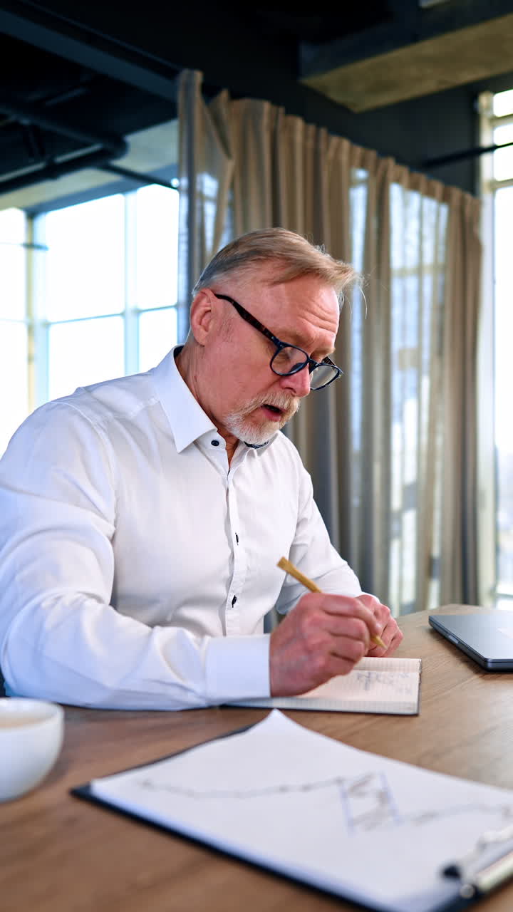 Bearded male in white shirt sits at desk writing. Businessman planning his day or taking notes about ideas or strategies. Vertical video.