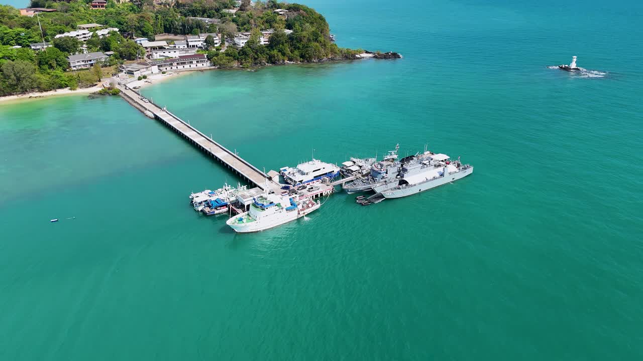 Aerial footage captures navy ships docked at a pier in Phuket, Thailand. Clear skies and turquoise waters create a serene atmosphere