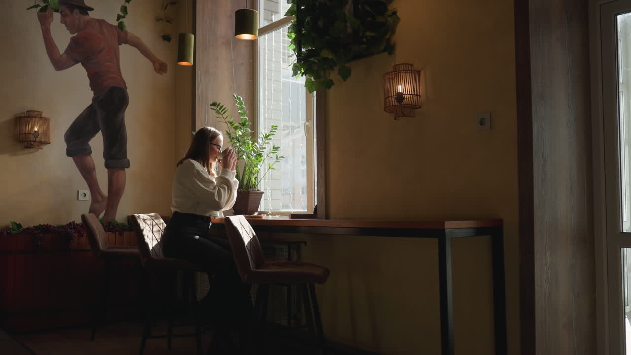 Elegant lady seated beside large window sips coffee peacefully as warm sunlight shines through, potted plant and expressive wall art create serene ambiance in cozy, stylish interior setting