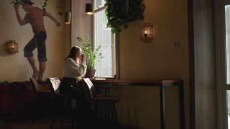 elegant lady seated beside large window sips coffee peacefully as warm sunlight shines through, potted plant and expressive wall art create serene ambiance in cozy, stylish interior setting