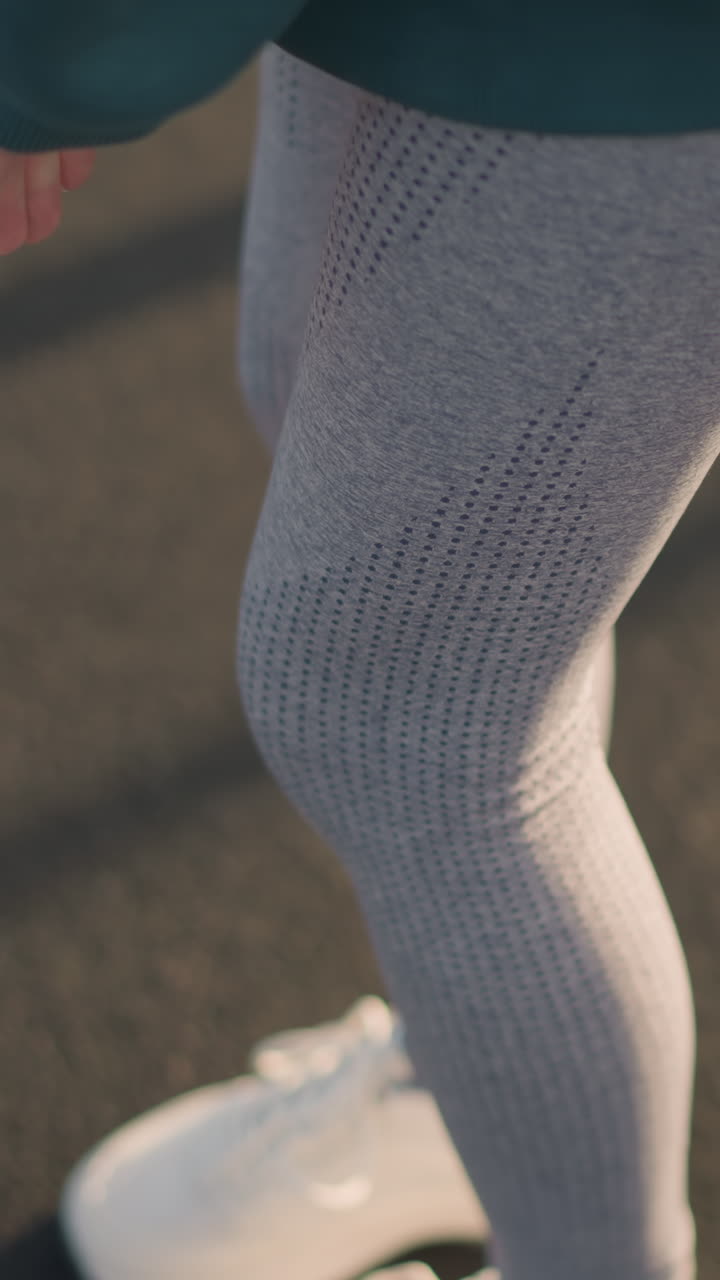 Primo piano di una donna con scarpe da ginnastica bianche in tela che fa stretching con la mano sul ginocchio, concentrandosi sulla postura atletica e sul movimento, mostrando uno stile di vita sano in un ambiente di allenamento all'aperto con ombre