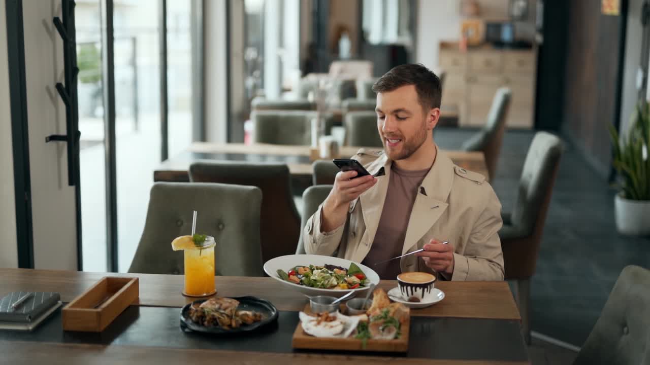 agradable blogger masculino adulto habla de comida en la cámara del teléfono inteligente sentado en la mesa del restaurante