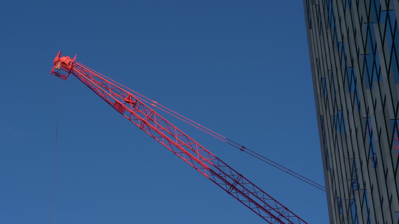 햇빛 아래 크레인의 붉은 팔이 마천루 창 앞 옆으로 움직인다