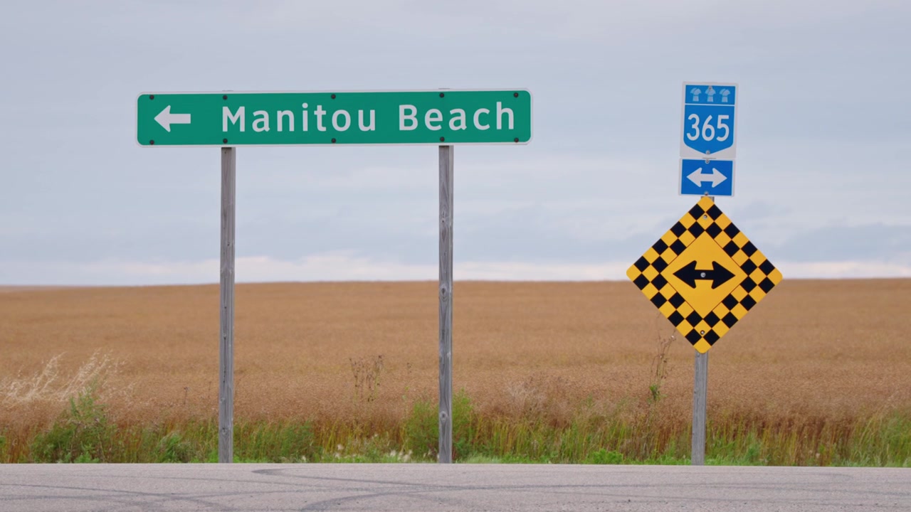 Statische Nahaufnahme des Straßenschildes von Manitou Beach, das nach links zeigt, mit einer Straße davor und einem gleichmäßigen goldenen Weizenfeld dahinter im ländlichen Saskatchewan, Kanada