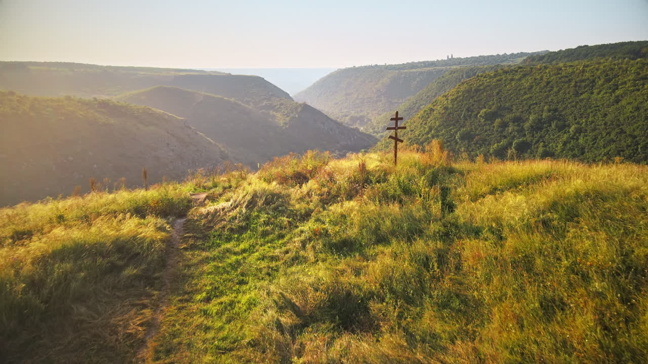 Aerial drone view of a valley with hill slopes covered with lush greenery, wooden cross on a hill's top on the foreground in Moldova