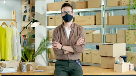 young caucasian man in mask looking at camera in good mood sitting on table in own shop with clothes