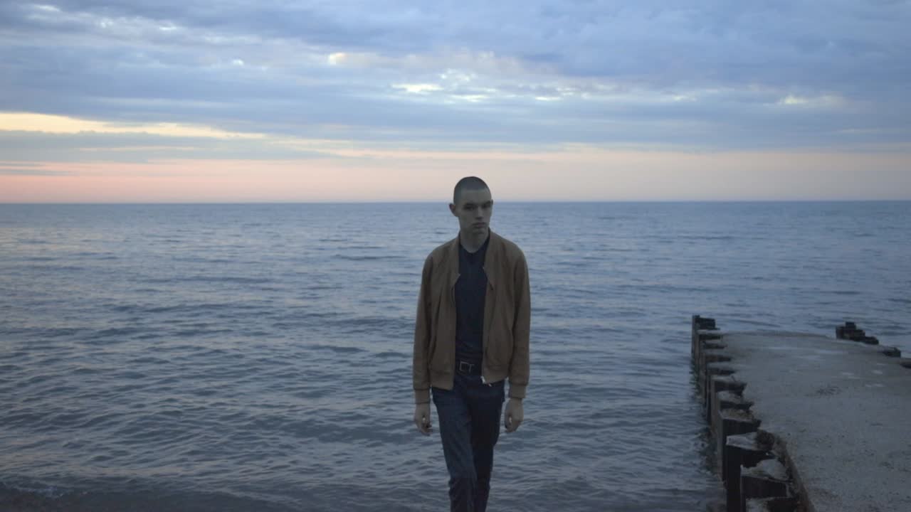 A Young Man Leaving The Shore Walking Towards The Camera With Blue Sea And Sky Background - Slow Motion