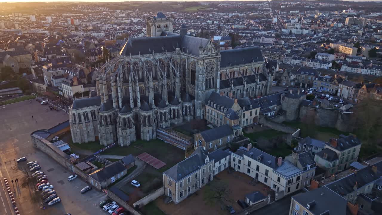 Ripresa con drone da un'angolazione verso il basso che si allontana dalla Cattedrale di Le Mans al tramonto, rivelando la piazza accanto e la città circostante immersa in una calda luce dorata. Francia