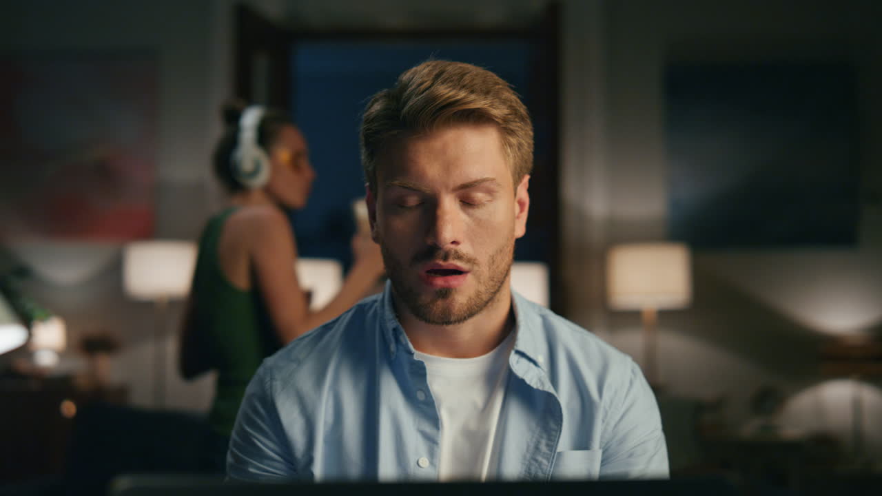 Thoughtful freelancer video chatting. Guy watching laptop while wife dancing