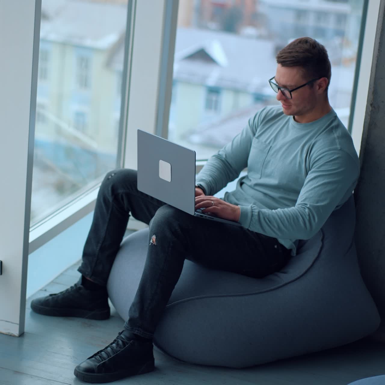 Comfortable work conditions in the modern office. Happy young man sitting in bean bag chair typing on his laptop at looking at window. High angle view
