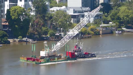 a small crane is transported up a river on a flat deck barge that is being manoeuvred by a small tugboat