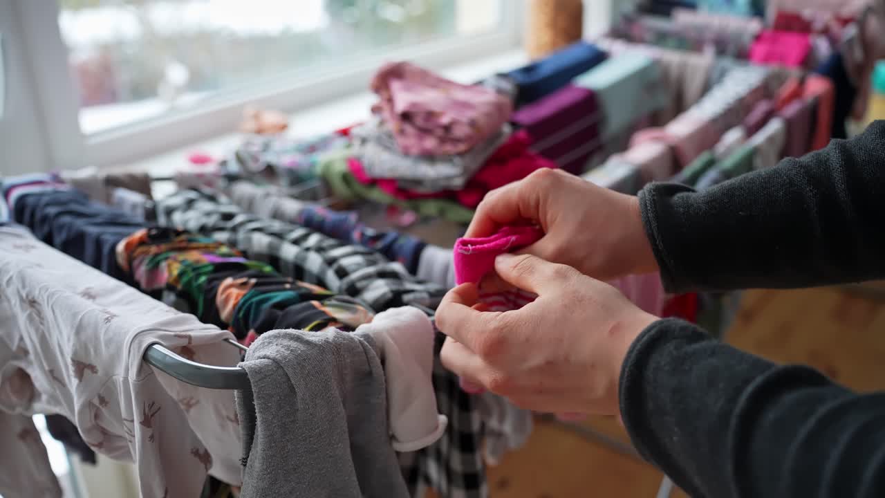Mani che rimuovono calzini rosa per neonati da uno stendibiancheria interno accanto a una finestra con biancheria pulita. Facende domestiche, organizzazione dei vestiti, routine familiare, riprese aggiuntive di vita domestica accogliente