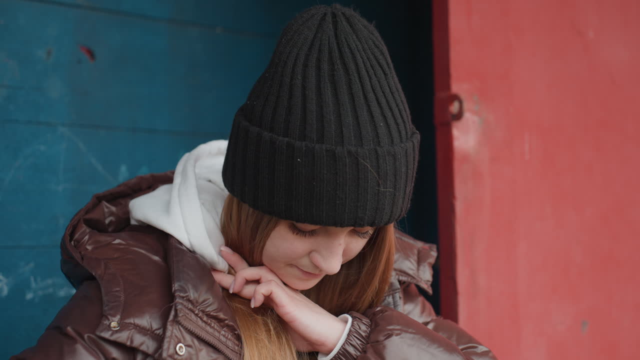 흰색 벽이 칠해진 문 앞에서 머리카락을 귀 뒤로 넘기고 검은색 비니와 광택 있는 패딩을 입고, 파란색과 빨간색 벽 텍스처를 배경으로 부드럽고 수줍은 미소를 짓고 있는 백인 여성, 친밀한 겨울 분위기와 부드러운 자연광이 담긴 솔직한 거리 사진