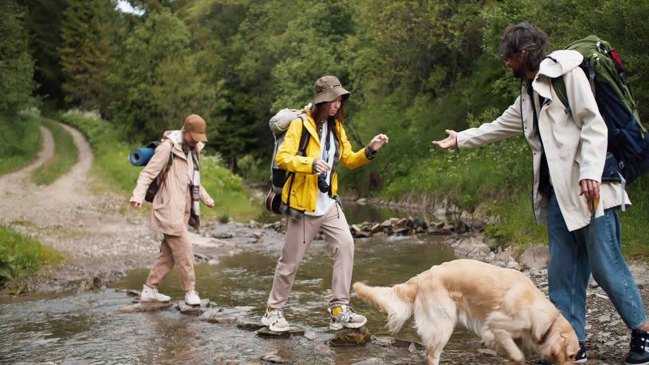 el tipo ayuda a dos chicas a cruzar un río de montaña en piedras especiales, le da una mano para que lo agarren y crucen al otro lado. personas con ropa de senderismo nuestra puerta
