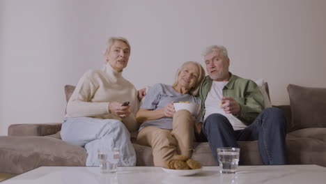 group of three senior people watching tv, talking and eating caramel popcorn while sitting on sofa at home 2