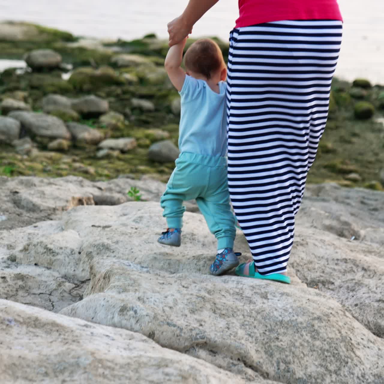 Lovely toddler boy in blue clothes stepping by the grey stones. Mom in long striped skirt supporting her child by the hands. Waterfront at backdrop