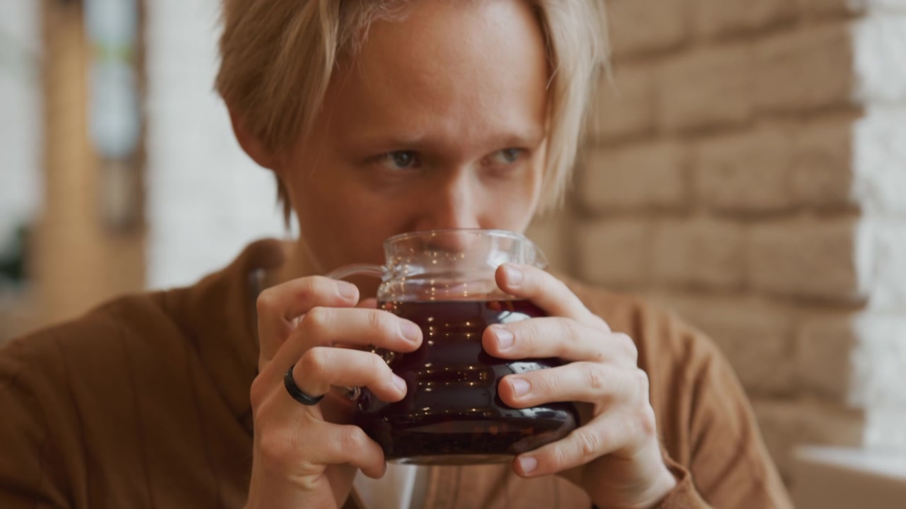 Primo piano di un uomo che tiene in mano e beve da una brocca di vetro piena di liquido scuro. Il soggetto indossa una giacca marrone, con un focus sulle mani che afferrano il manico mentre beve in un ambiente interno.