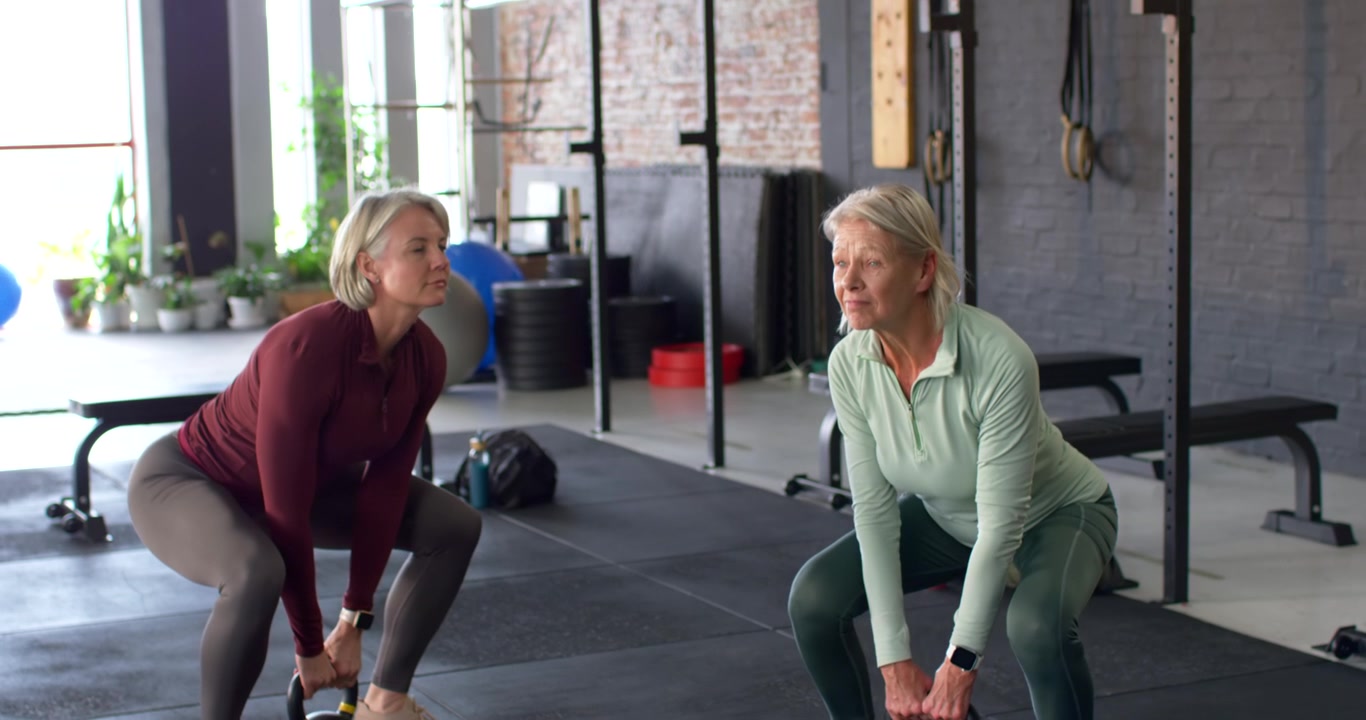 Due donne che sollevano kettlebell piegando fianchi e ginocchia fino a una posizione eretta in palestra per la forza