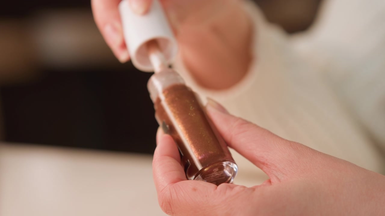 Macro primo piano di mani che sollevano e ruotano un applicatore a pennello da una piccola bottiglia lucida con tappo bianco, rivelando un liquido scintillante color bronzo-oro che aderisce, si allunga e forma goccioline a.