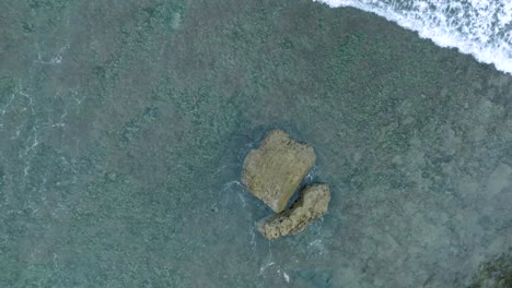 Top-Down-Slow-Motion-Drone-View-of-Waves-crashing-over-shallow-coral-reef-and-big-rock-in-Uluwatu-Bali-Indonesia