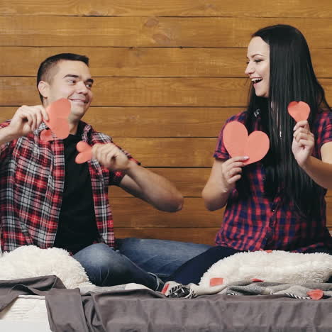 romantic couple sitting on bed holding red paper hearts in both hands make movements by hands. happy man shows motion to different sides of hands and his girl repeats after him.
