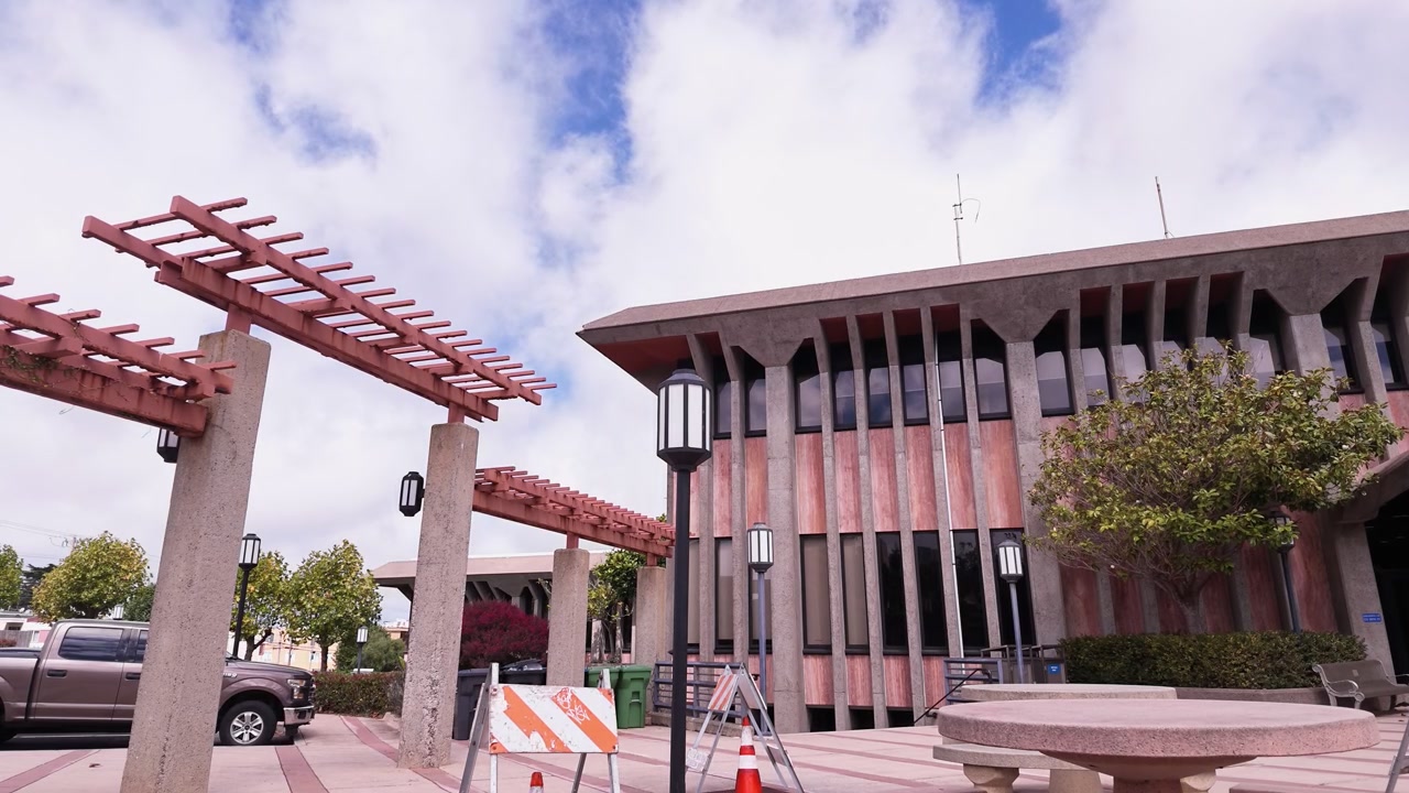 Arquitetura modernista do edifício da Prefeitura em Daly City, Califórnia, exibindo sua fachada de concreto distinta sob um céu azul com nuvens brancas suaves, um cenário governamental urbano tranquilo