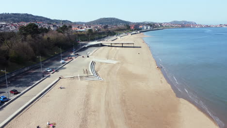 An-aerial-view-of-Colwyn-Bay-seafront-on-a-sunny-day,-flying-over-the-beach-towards-Rhos-on-Sea,-Denbighshire,-North-Wales,-UK