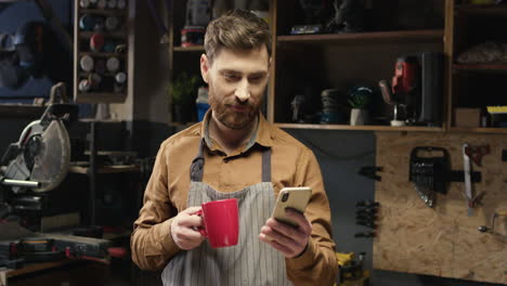 caucasian young male carpenter in apron resting in workshop, sipping hot drink and using smartphone