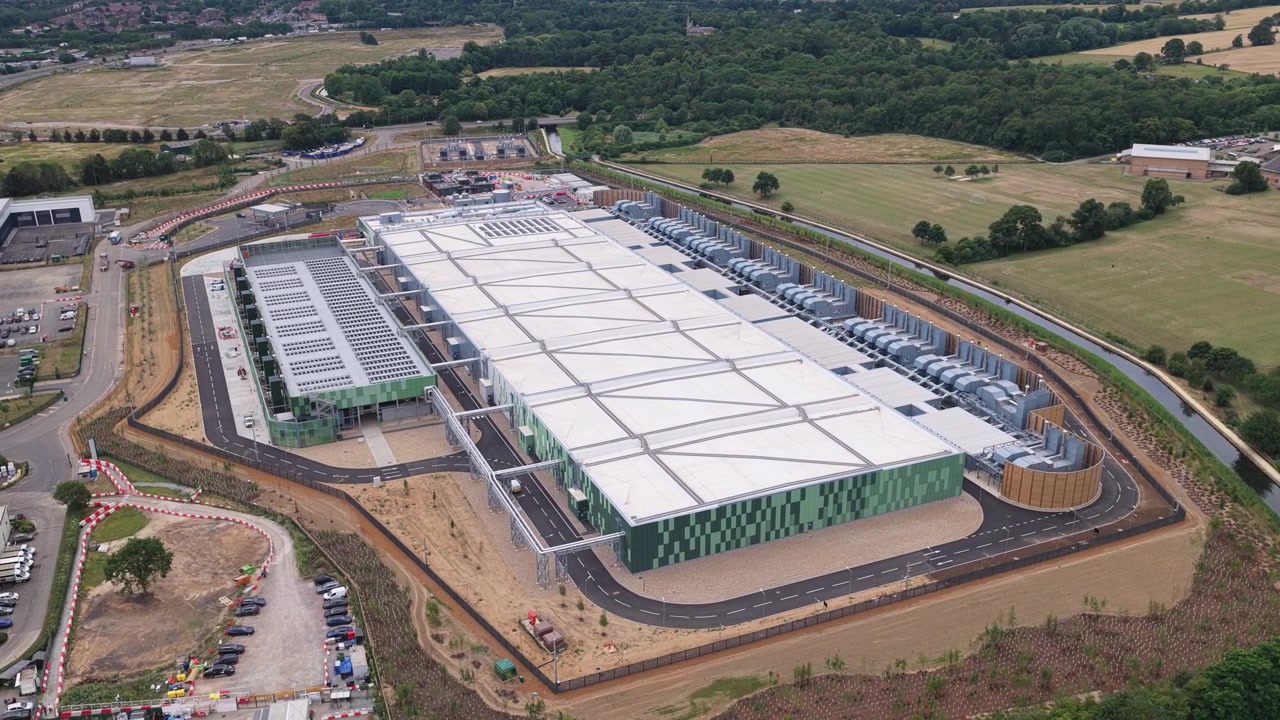 Grand complexe de centre de données industriel entouré de champs verts pendant la journée avec une visibilité claire des infrastructures et des aires de stationnement pour des opérations sécurisées et des tâches de maintenance.
