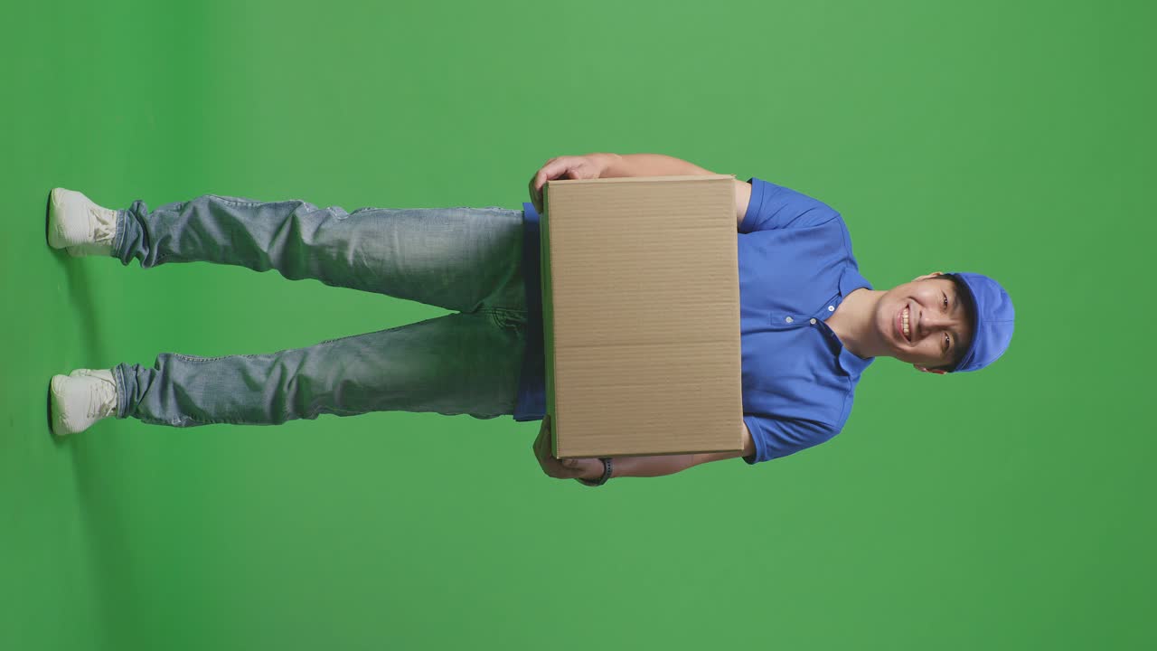 Full Body Of Asian Male Courier In Blue Uniform Smiling While Delivering A Carton On Green Screen Background In The Studio