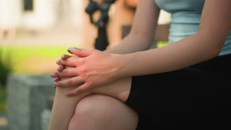 partial view of young woman sitting outdoors with legs crossed, hands clasped on knee making gentle movement with thumbs, with warm sunlight illuminating her skin with blurred background